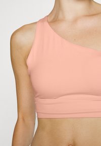 Top desportivo curto em rosa claro, com design de um ombro, textura suave e silhueta ajustada. Apresenta uma banda lisa na bainha.