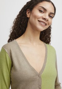 Cardigan a V con maniche lunghe, caratterizzato da un lato beige e dall'altro verde, realizzato con tre bottoni e una texture morbida.
