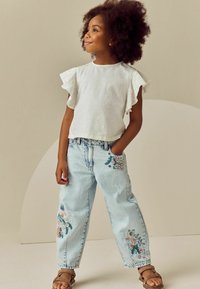 Witte top met korte mouwen en kantafwerking, lichtblauwe, versleten denim jeans met kleurrijke bloemenborduursel en tan sandalen.