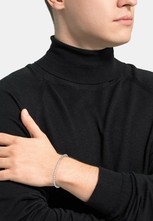 Pulsera de plata trenzada llevada en una mano, con un suéter de cuello alto negro y acanalado. La pulsera tiene una forma circular y una textura suave.