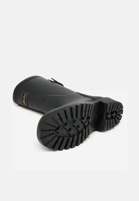 Must värvi kummist vihmakepid tekstureeritud tallaga, ümmarguse varbaga, silmapaistva tallapatruni mustriga ja väike embleem ülemise osa lähedal. Küljel on ka pandlakaunistus.