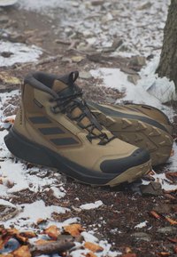 Hellbraune und schwarze Wanderschuhe auf einem verschneiten Waldboden mit robusten Sohlen und Schnürsenkeln, in der Nähe eines Baumstamms und verstreuter Blätter.