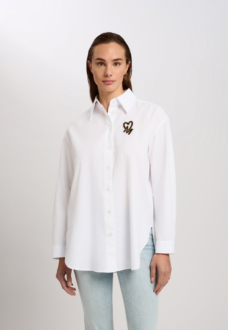 White button-up shirt with a classic collar, featuring a black and gold embroidered heart and "M" detail on the left chest. Smooth texture.
