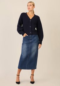 Marineblaue gerippte Strickjacke mit V-Ausschnitt und drei Knöpfen, kombiniert mit einem Jeans-Maxikleid mit kleinen Perforationen, ergänzt durch schwarze Absätze.