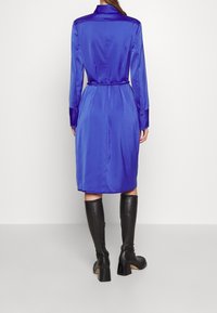 Modrá saténová košilová šaty s dlouhými rukávy a opaskem v pase, s klasickým límcem a knoflíkovými manžetami, doplněné nadkolenními černými botami.