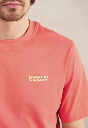 Mann trägt ein korallenrotes T-Shirt mit kurzen Ärmeln und dem Aufdruck "AMALFI COAST" auf der linken Brust vor einfarbigem Hintergrund.