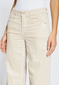 Beige corduroy pants with vertical ribbing, featuring a button closure, two front pockets, and a logo patch on the waistband.