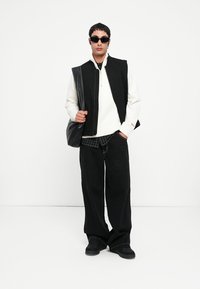 Vest negru peste un pulover alb cu fermoar, stratificat cu o cămașă cu carouri. Pantaloni negri lejeri și încălțăminte neagră completează ținuta. Ochelari de soare închiși la culoare purtați.