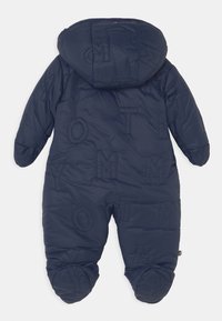 Laivastonsininen paksu lumipuku hupulla, jossa on kohokirjaimia, tilkkutyylinen pinta ja joustavat hihansuut nilkoissa ja ranteissa.