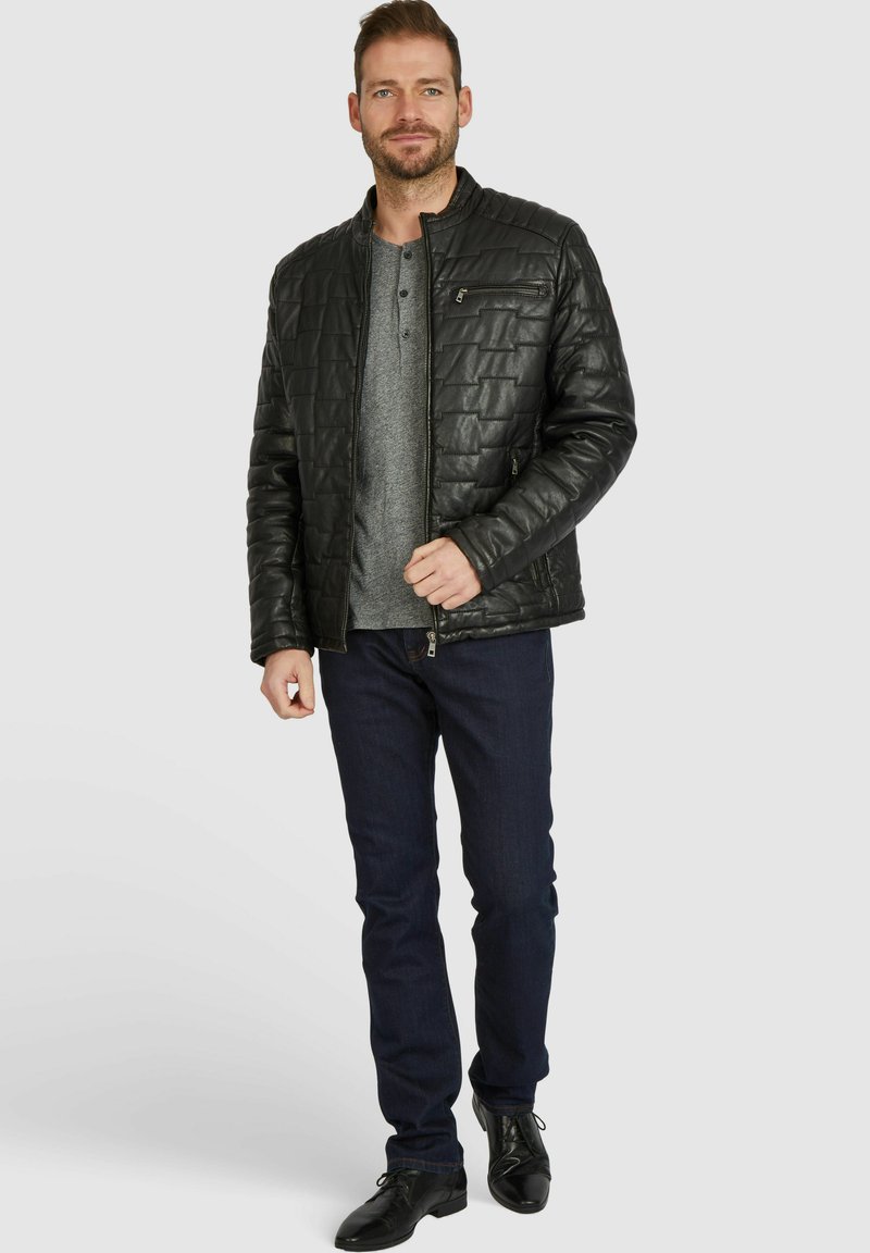 Svart vadderad jacka med dragkedja framtill och ståkrage, buren över en grå henley-tröja och mörka jeans. Slät lädertextur, avslappnad passform.