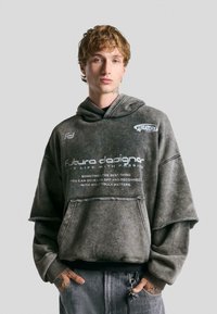 Oversized, gray hoodie with a faded texture and frayed hem. Features text print and two logos on the front, with a kangaroo pocket.