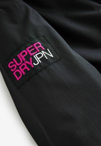 Fekete anyag, függőleges bordázott textúrával, amelyen egy téglalap alakú folt található. A folton pink és fehér hímzéssel a "SUPER DRY" felirat szerepel.