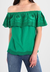 Zelený top bez ramínek s rozšířenou vrstvou, který má výřez na lemu a texturovanou látku, který je zkombinován s modrými džíny.