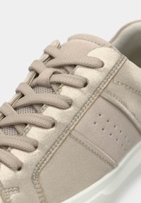 Μπεζ αθλητικά παπούτσια με λεία υφή, στρογγυλεμένη μύτη και διακοσμητική διάτρηση στο πλάι. Τα κορδόνια είναι υφασμένα, με μια διακριτική λάμψη στο επάνω υλικό.