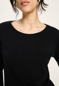 Frau in einem schwarzen Rundhals-Langarmshirt, das von den Schultern bis zum oberen Brustbereich sichtbar ist, mit Haaren, die lose das Gesicht umrahmen.