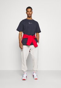 Camiseta de algodón azul marino con logo, pantalones de chándal blancos y sudadera roja atada a la cintura, combinados con zapatillas blancas y rojas.