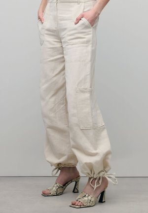 Persoon die beige cargobroek draagt met trekkoord aan de pijpen en open teen schoenen met hoge hakken en slangenhuidpatroon, staand tegen een effen achtergrond.