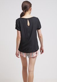 Donna con i capelli castani raccolti in una coda di cavallo bassa, che indossa una maglietta nera a maniche corte con un'apertura a chiave sul retro e pantaloni corti a fantasia, in piedi davanti a uno sfondo neutro.