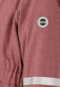 Röd, texturerad tygjacka med en cirkulär logopatch och en reflekterande silverrand på ärmen.