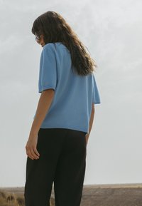 Gaiši zils īsām piedurknēm džemperis ar brīvu piegriezumu, izgatavots no mīksta adījuma materiāla. Apvienots ar melniem platām biksēm, redzams no aizmugures.