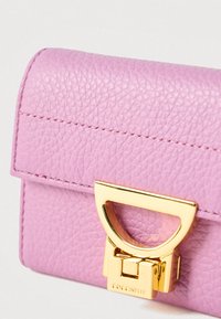 Levandų tekstūruotas odinis piniginė su aukso spalvos metaliniu užsegimu, ant kurio išgraviruotas prekės ženklas, parodyta prieš baltą foną.