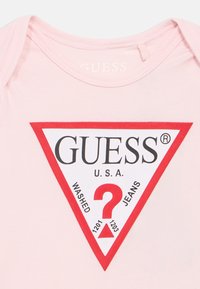 Ροζ βαμβακερό κορμάκι με εμφανές κόκκινο και λευκό τριγωνικό λογότυπο με τη φράση «GUESS U.S.A.» και ένα σχέδιο με ερωτηματικό.