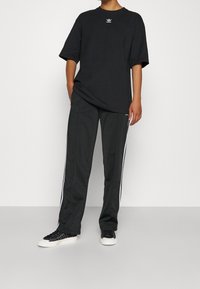 Black cotton oversized t-shirt with white Adidas logo, paired with dark gray track pants featuring white side stripes and black sneakers.