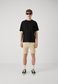 Sort kortærmet t-shirt med en frontlomme, beige shorts, sorte sneakers med hvide såler og solbriller. Minimalistisk design, afslappet pasform.