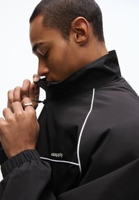 Black windbreaker jacket with white accents, high collar, and elastic cuffs. Features a zip closure and a small logo on the chest.