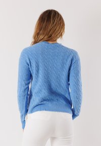 Donna con lunghi capelli castani, che indossa un maglione azzurro chiaro a fantasia e pantaloni bianchi, rivolta di spalle su uno sfondo chiaro e semplice.