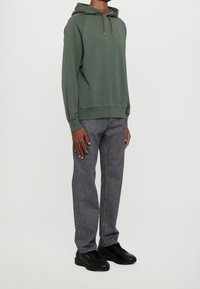 Felpa verde con tasca a marsupio e cordino, abbinata a jeans grigi e sneakers nere. Design casual e rilassato, materiale in cotone misto.