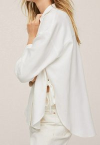 Weiße langärmlige Bluse mit lässigem Schnitt, ausgestattet mit seitlichen Bindebändern, einem abgerundeten Saum und leichtem Stoff, getragen über hellen Hosen.