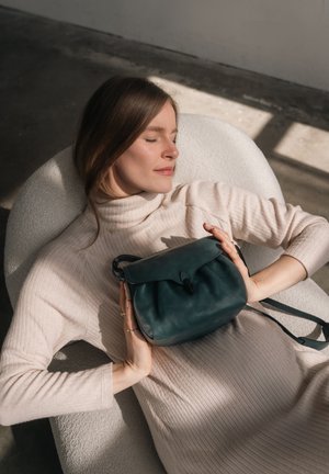 Borsa a tracolla in pelle verde acqua con chiusura a pattina, forma arrotondata e tracolla regolabile, tenuta da una persona in un maglione beige a coste.