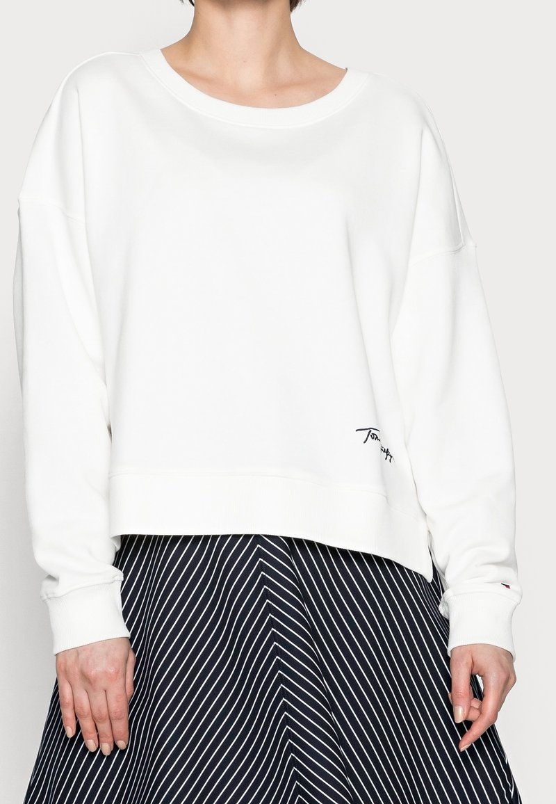 Sweat-shirt court blanc à large encolure et manches longues, avec un logo brodé près de l'ourlet, accompagné d'une jupe rayée marine.