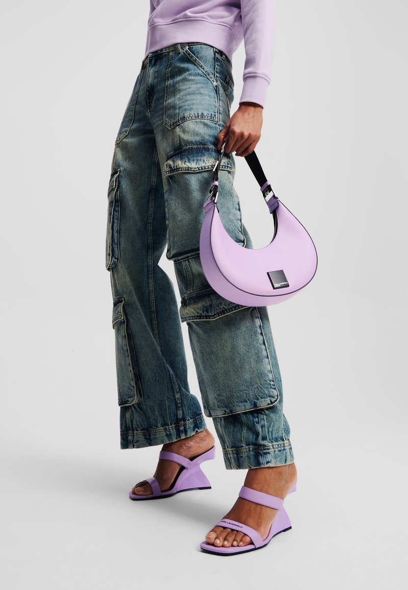 Bolsa lilás em forma de crescente com alça preta, combinada com calças cargo de denim azul de corte solto e sandálias lilás de salto.