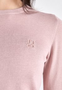 Zbliżenie osoby w jasnoróżowym swetrze z dzianiny z małym haftowanym logo "HUGO" na piersi.