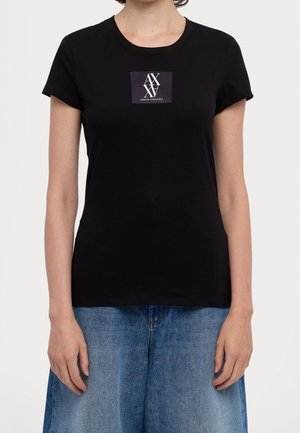 Nainen pukeutunut istuvaan mustaan lyhythihaiseen T-paitaan, jossa on harmaa neliskulmainen "AX Armani Exchange" -logo rinnassa, yhdistettynä sinisiin farkkuihin.