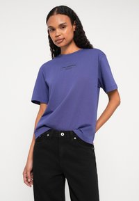 Fialové bavlněné tričko s krátkým rukávem, se tónovým potiskem loga "KARL LAGERFELD JEANS" na hrudi. Nosené s černými denimovými kalhotami.