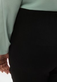 Μαύρο παντελόνι με λεία υφή, συνδυασμένο με ένα ανοιχτό πράσινο, μακρυμάνικο τοπ με υφή. Έμφαση στο ύφασμα και την αντίθεση χρωμάτων.