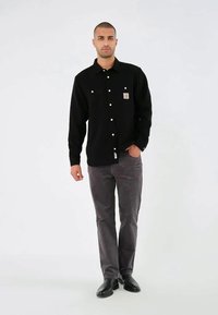 Black long-sleeve shirt with button closure and chest pocket. Worn with gray corduroy pants and black shoes. Simple, clean design.