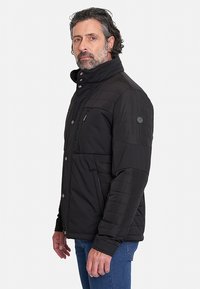 Chaqueta aislante negra con un cuello alto, cierres a presión y cremallera, paneles acolchados y bolsillos laterales, llevada sobre unos vaqueros azules.