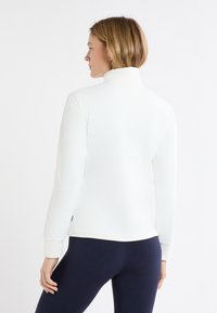 Biała kurtka z długim rękawem z wysokim kołnierzem i mankietami, gładka w dotyku, zestawiona z ciemnymi legginsami. Brak widocznych logo i wzorów.