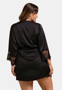 Robe en satin noir avec des manches trois quarts présentant des accents en dentelle transparente aux poignets et une taille nouée. Texture lisse avec une coupe décontractée.