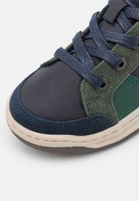 Zapatilla con puntera de cuero azul marino, acentos de ante verde, panel de malla texturizada y cordones azul marino. Presenta una suela de goma con un ligero borde beige.