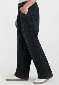 Zwarte wijde jeans van textuurstof, met zij- en achterzakken. Gepaard met witte sneakers voor een casual look.
