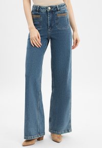 Vrouw in een hoge taille, wijde blauwe jeans met voorzakken en beige puntige schoenen, staande op een witte achtergrond.