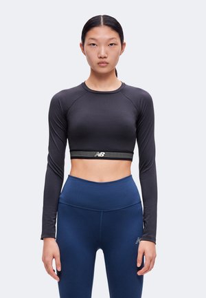 Kvinde iført en sort langærmet crop top og blå leggings med høj talje stående mod en ensfarvet lys baggrund.