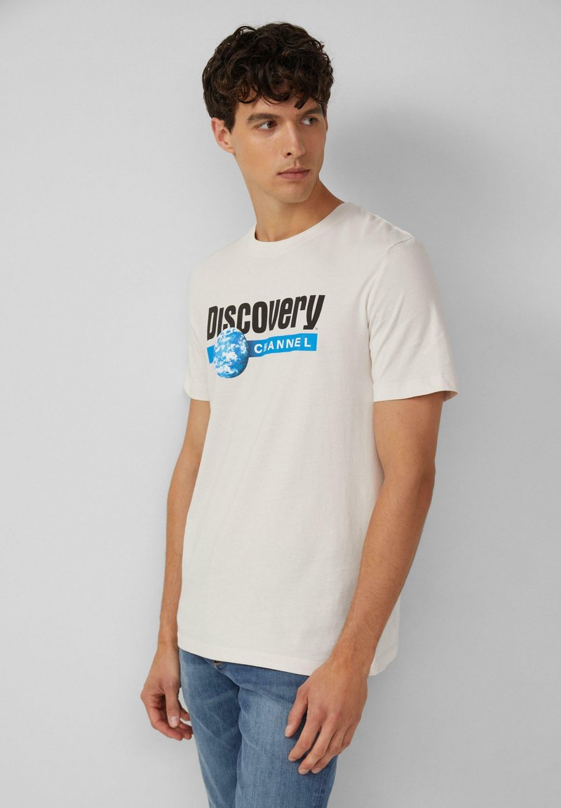 Jasno beżowa bawełniana koszulka z krótkim rękawem, z napisem "Discovery Channel" oraz niebieską grafiką globu na przodzie.