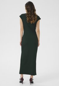 Robe vert foncé, longueur cheville, à manches courtes et silhouette ajustée. Tissu lisse avec un discret détail sur les côtés. Portée avec des talons hauts bordeaux.
