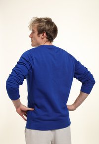 Modrá mikina s žebrovanými manžetami a lemem, s kulatým výstřihem a měkkou, texturovanou tkaninou. Pohled zezadu, ruce na bocích.